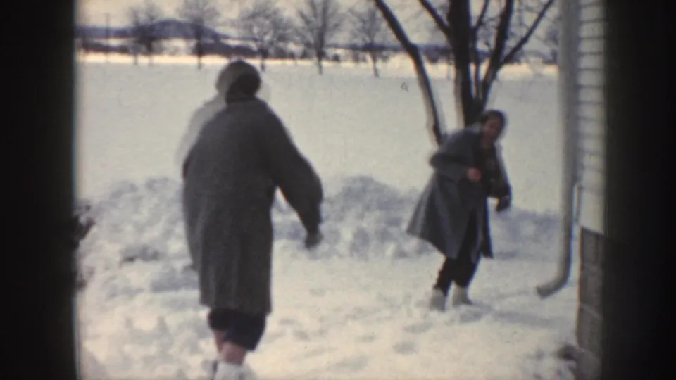 Two adults walk through a snowy Nebraska residential yard, with house siding, bare trees, and open winter fields beyond.