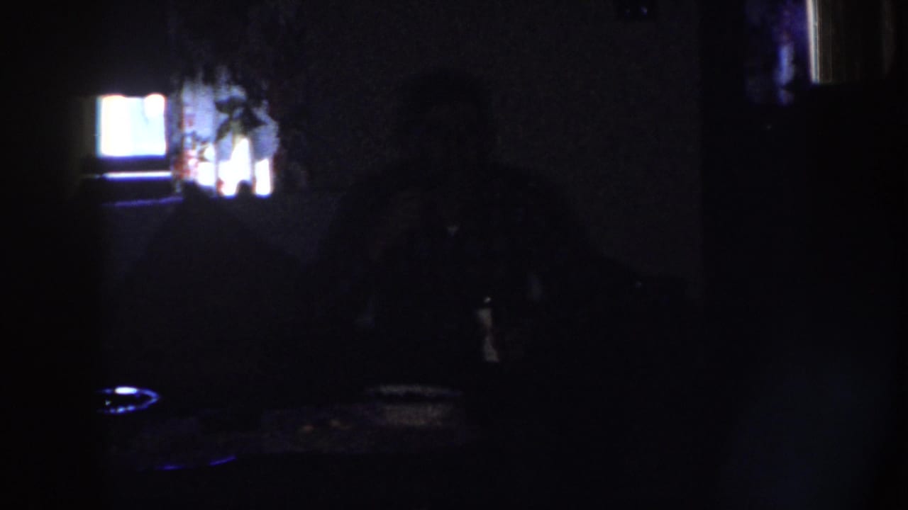 1958 Nebraska 8mm frame showing a dim private interior with a silhouetted seated adult, tabletop, and bright rear window or screen.