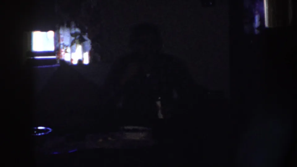 1958 Nebraska 8mm frame showing a dim private interior with a silhouetted seated adult, tabletop, and bright rear window or screen.