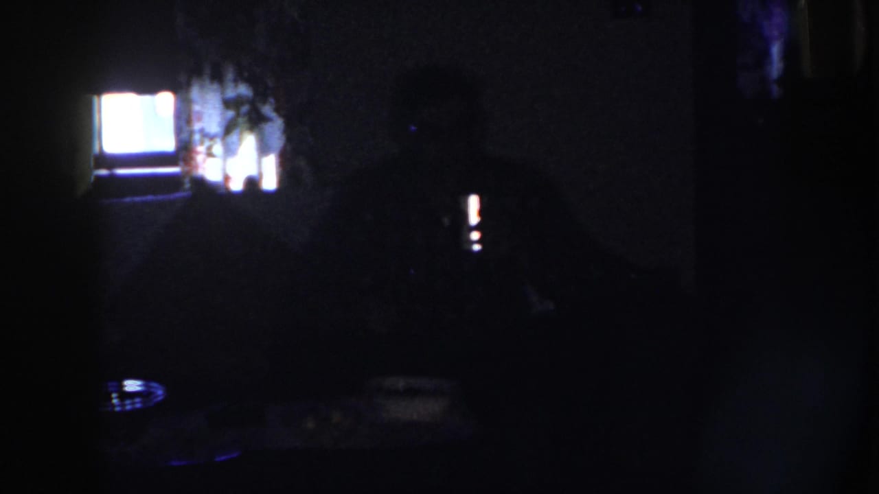 1958 Nebraska home-movie frame with a dark seated figure at a table, a bright rear window or screen, and faint room details.