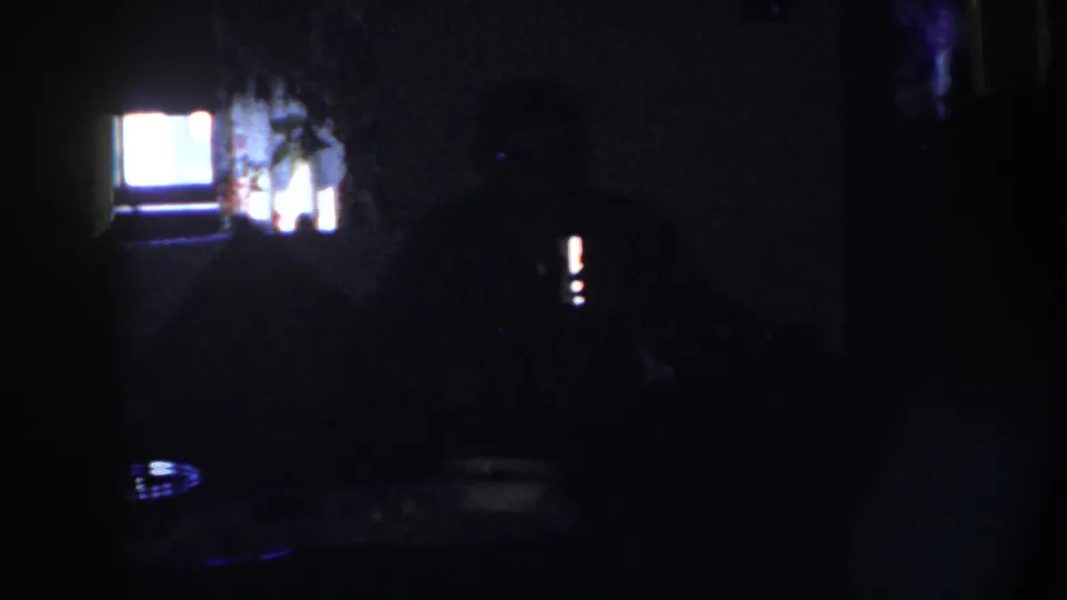 1958 Nebraska home-movie frame with a dark seated figure at a table, a bright rear window or screen, and faint room details.