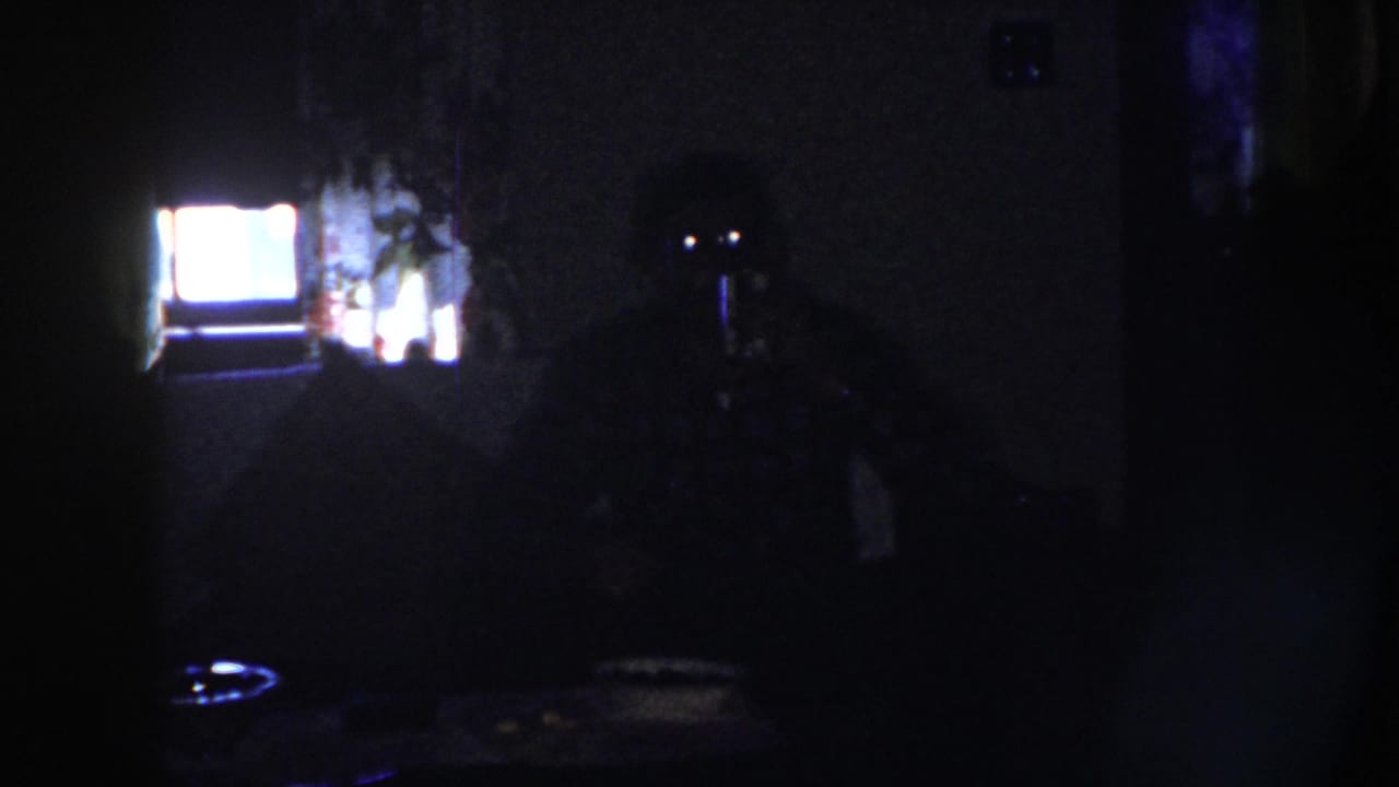 Dark 1958 Nebraska home-movie frame showing a dim room with a glowing television screen and indistinct household shapes.