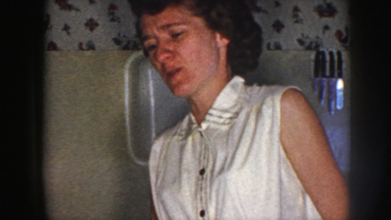 1958 Nebraska home-movie frame of a woman in a white sleeveless bow-collar blouse in a kitchen with floral wallpaper.