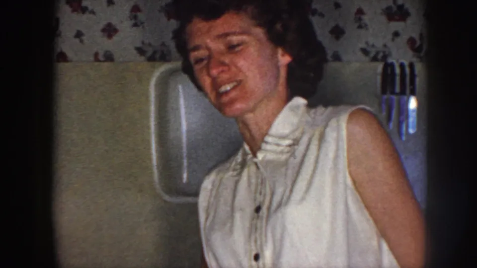 1958 Nebraska home-movie frame showing the woman smiling in a kitchen, with floral wallpaper and wall-mounted kitchen items nearby.