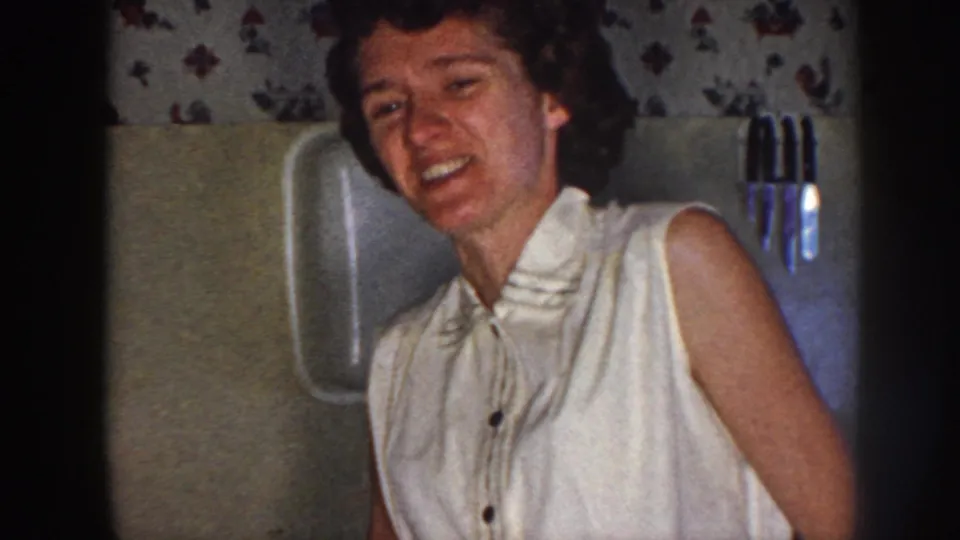 1958 Nebraska home-movie frame of the woman laughing or smiling toward the camera in a modest kitchen interior.