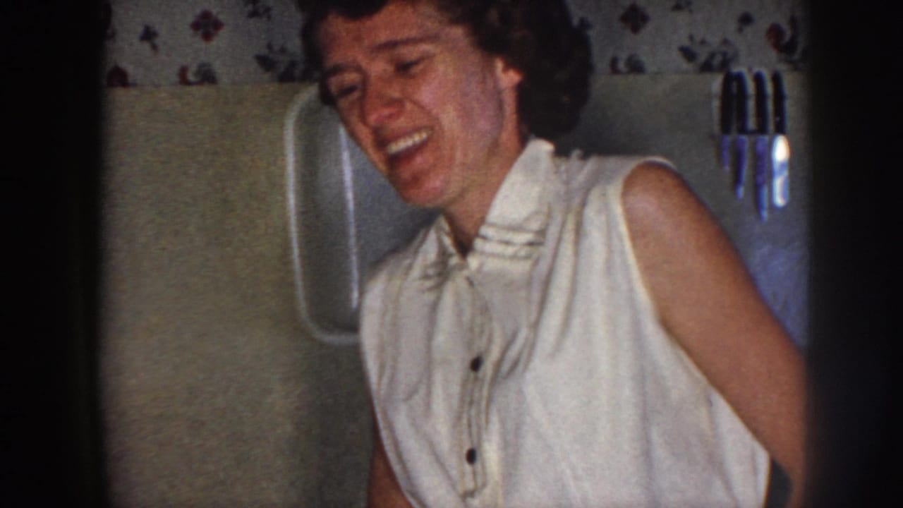 In 1958 Nebraska home-movie footage, a young woman in a light sleeveless blouse laughs in a softly lit private interior.