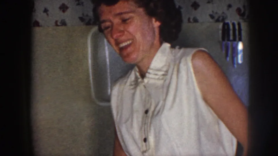 In 1958 Nebraska home-movie footage, a young woman in a light sleeveless blouse laughs in a softly lit private interior.
