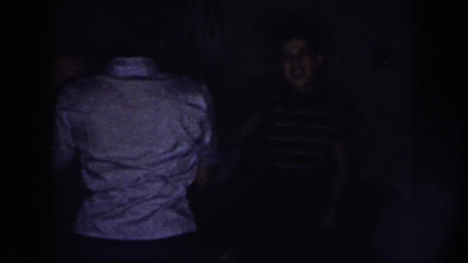 Two young women appear in a dim Nebraska home interior, one turned away while the other is partly visible in a striped shirt.