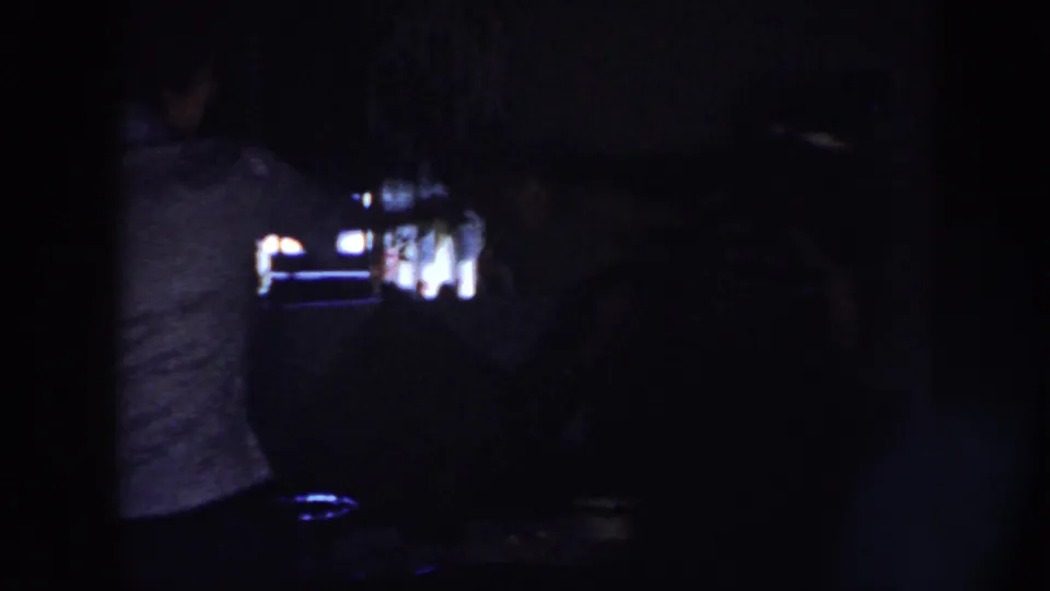 A dim 1958 Nebraska home-movie frame shows a woman from behind near small bright highlights in a private indoor space.