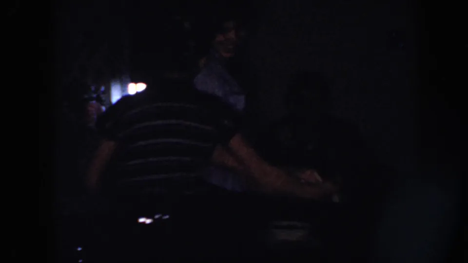 In a low-lit Nebraska home interior, a woman in a striped shirt leans or turns as another figure is faintly visible nearby.