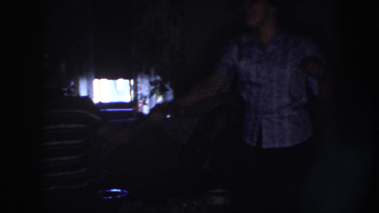 Two women dance in a dim Nebraska interior in 1958, one in stripes and one in a light patterned blouse, backlit by a bright screen.