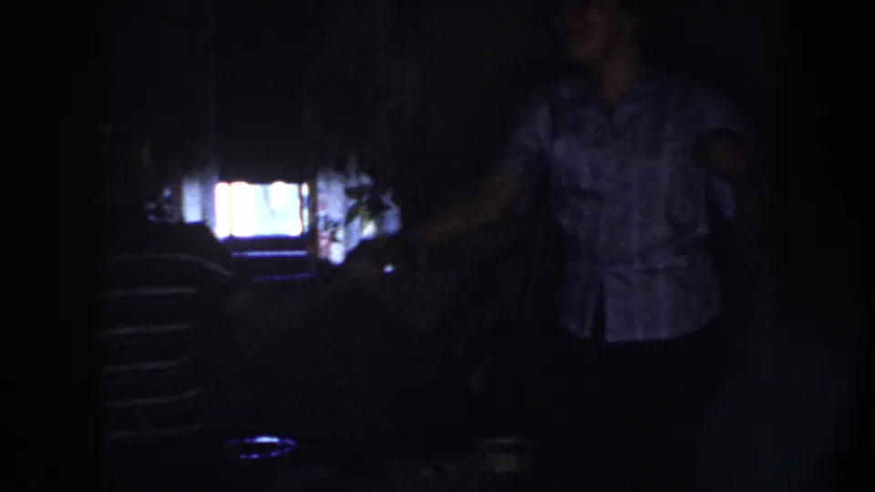Two women dance in a dim Nebraska interior in 1958, one in stripes and one in a light patterned blouse, backlit by a bright screen.