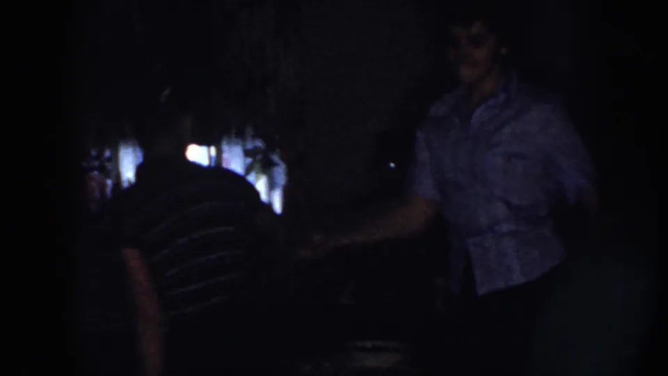 In this 1958 Nebraska home-movie frame, a woman in a patterned blouse gestures while another in a striped shirt stands partly silhouetted.