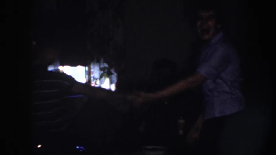 A woman in a light patterned blouse extends her arm toward another dancer in a dark Nebraska room, with bright backlight at left.