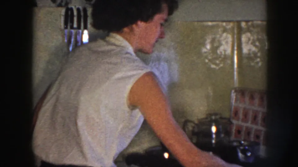 Nebraska 1958 kitchen view with a woman in a sleeveless white top leaning toward the sink or counter.