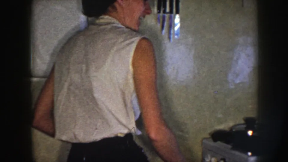 Nebraska 1958 home interior with a woman turned away in a tiled kitchen near cookware and a wall knife rack.