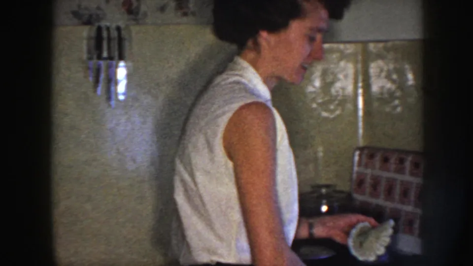 Nebraska 1958 kitchen frame showing a woman smiling at the counter with tiled walls, cookware, and patterned surface nearby.