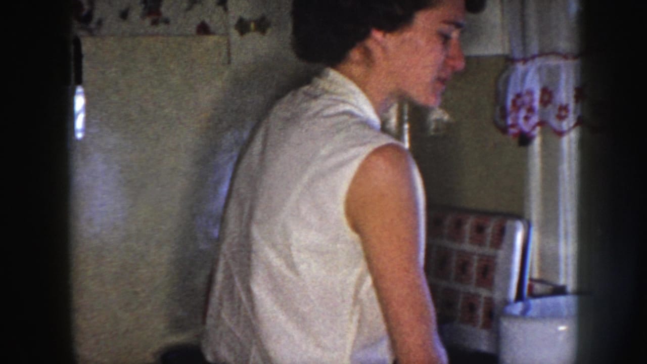 In a 1958 Nebraska home-movie frame, a woman stands side-on in a kitchen near a sink, tiled wall, window curtain, and counter items.