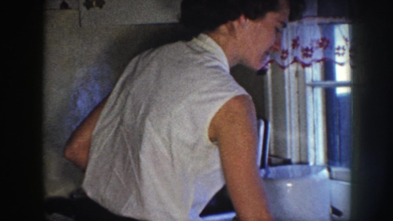 Woman leans forward at the sink near a window with a patterned curtain and tiled wall.