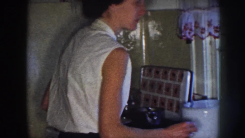 A 1958 Nebraska kitchen frame shows the woman from behind at the sink, with tile, curtain, counter objects, and home-movie film edges visible.
