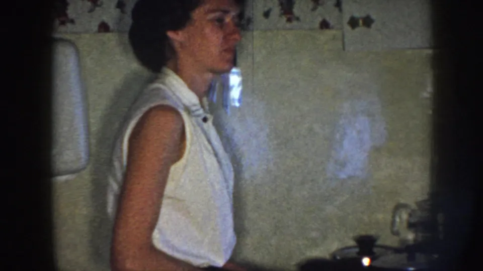 The woman turns slightly in profile in a 1958 Nebraska kitchen, with tiled wall, counter fixtures, and dark 8mm frame edges in view.