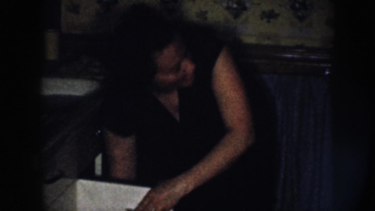 In a dim 1958 Nebraska home-movie frame, a woman leans over a kitchen sink with floral wallpaper and a radiator behind her.