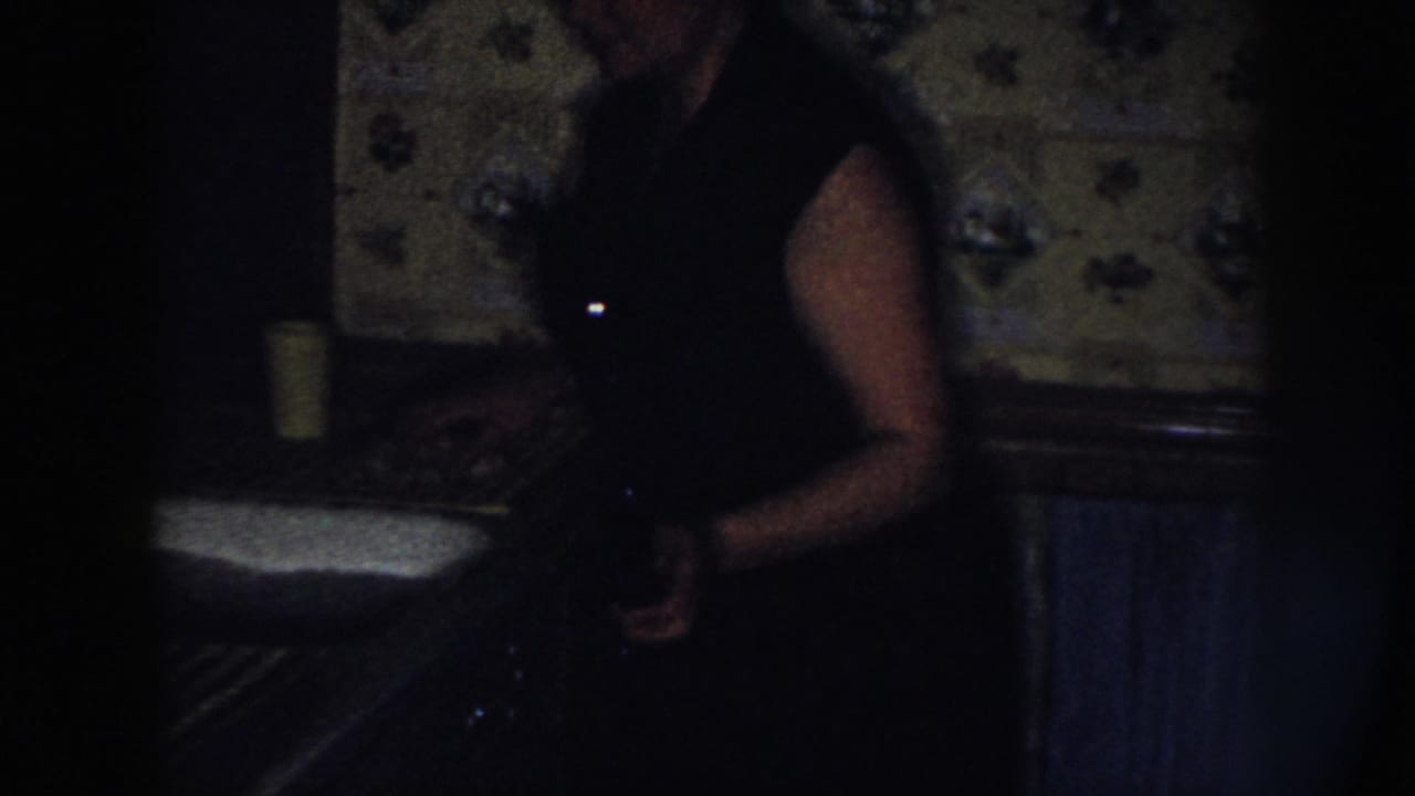 A woman stands beside the sink in a dark Nebraska kitchen interior, with floral wallpaper, radiator, and a yellow cup visible.