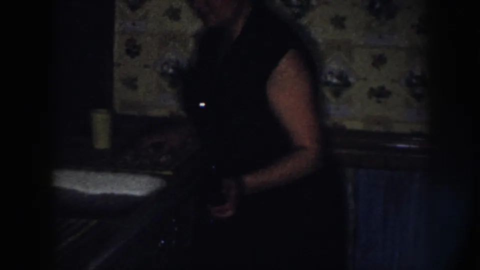 A woman stands beside the sink in a dark Nebraska kitchen interior, with floral wallpaper, radiator, and a yellow cup visible.