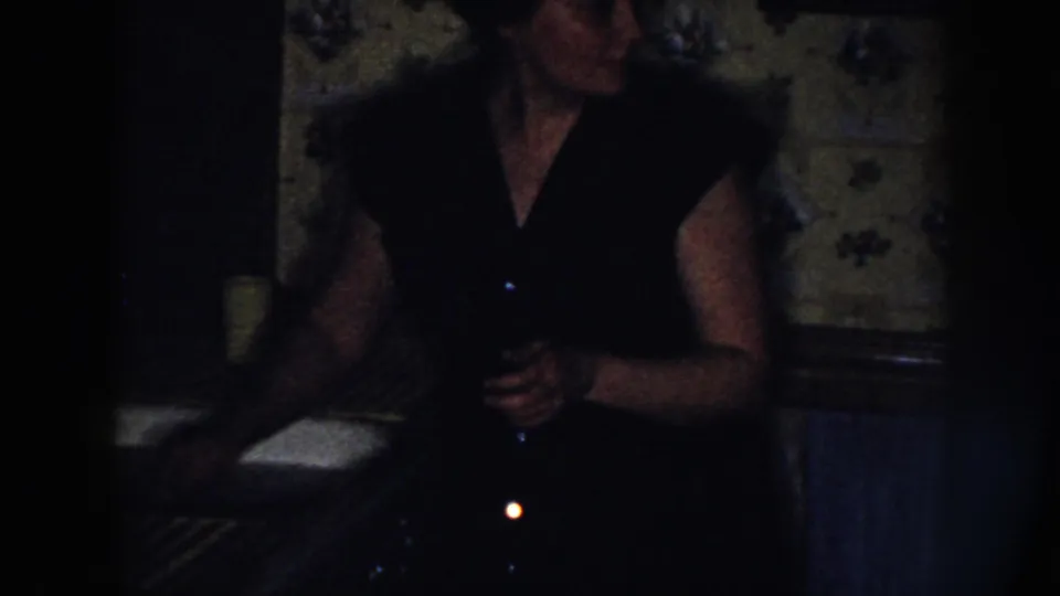 The woman turns beside the sink in the 1958 Nebraska kitchen scene, holding a dark object against a low-light background.