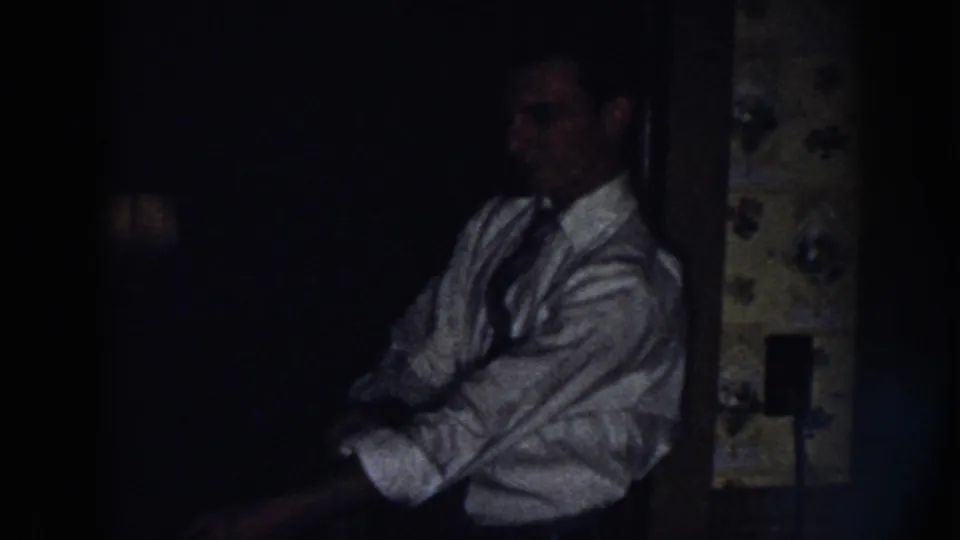 A man in a light shirt and tie sits near the kitchen area in the dim 1958 Nebraska home-movie scene.