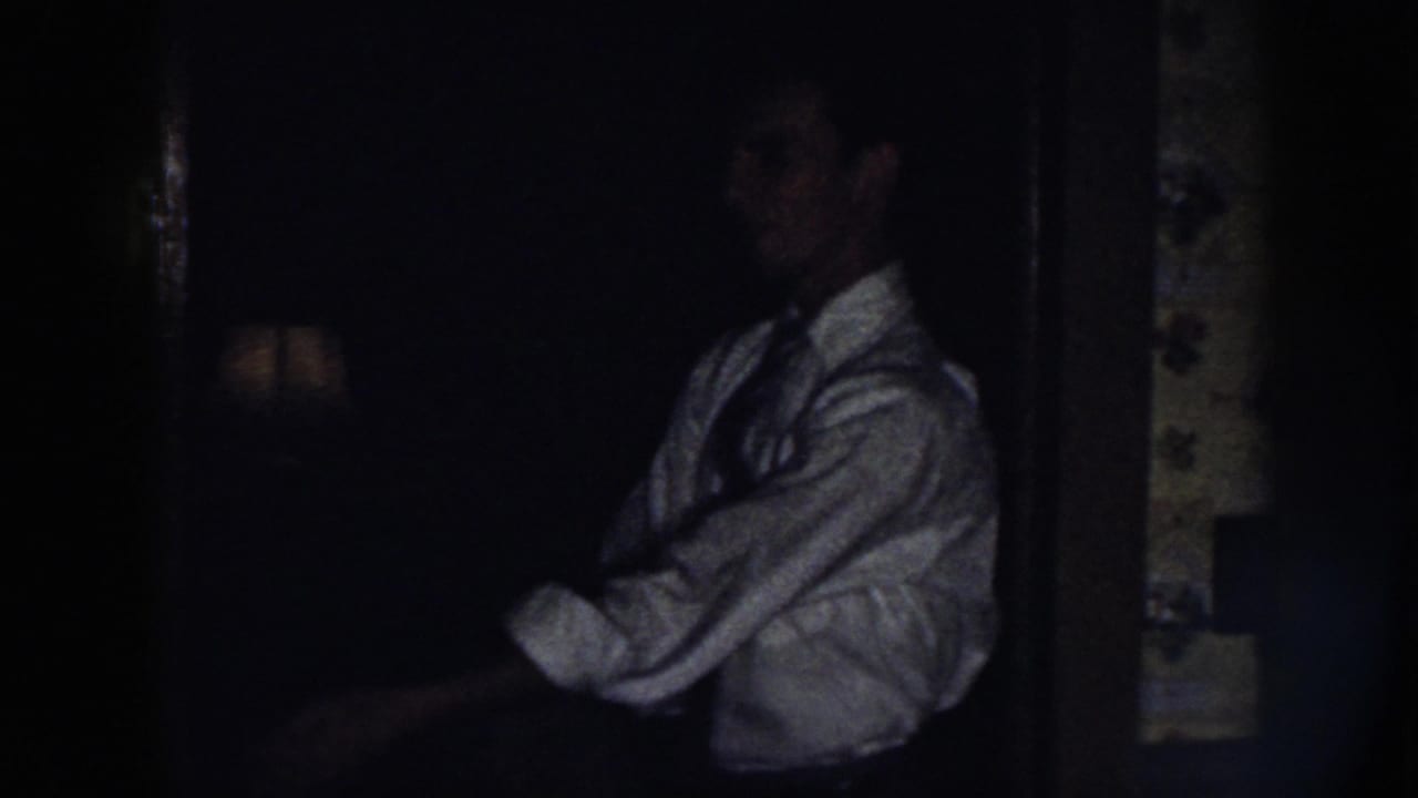 In a dim 1958 Nebraska home-movie frame, a man in a light dress shirt and dark tie adjusts his sleeve near a dark doorway.