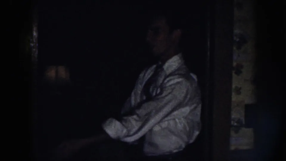 In a dim 1958 Nebraska home-movie frame, a man in a light dress shirt and dark tie adjusts his sleeve near a dark doorway.