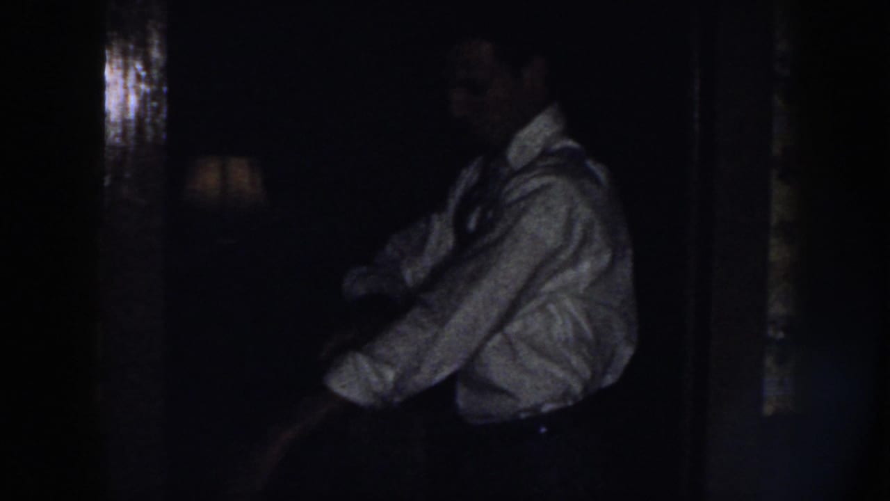 The man appears in profile inside a dim private interior, wearing a light shirt and tie while his sleeves are being adjusted.