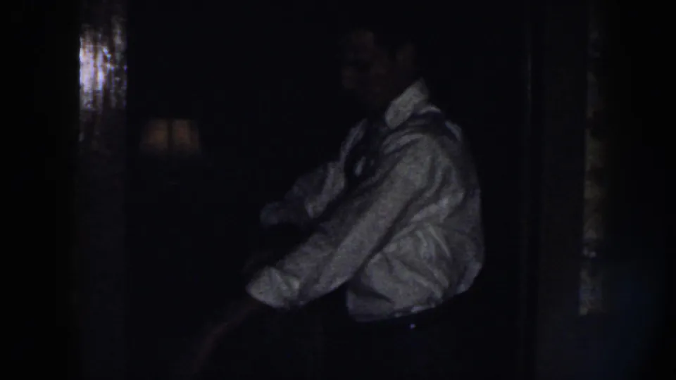 The man appears in profile inside a dim private interior, wearing a light shirt and tie while his sleeves are being adjusted.