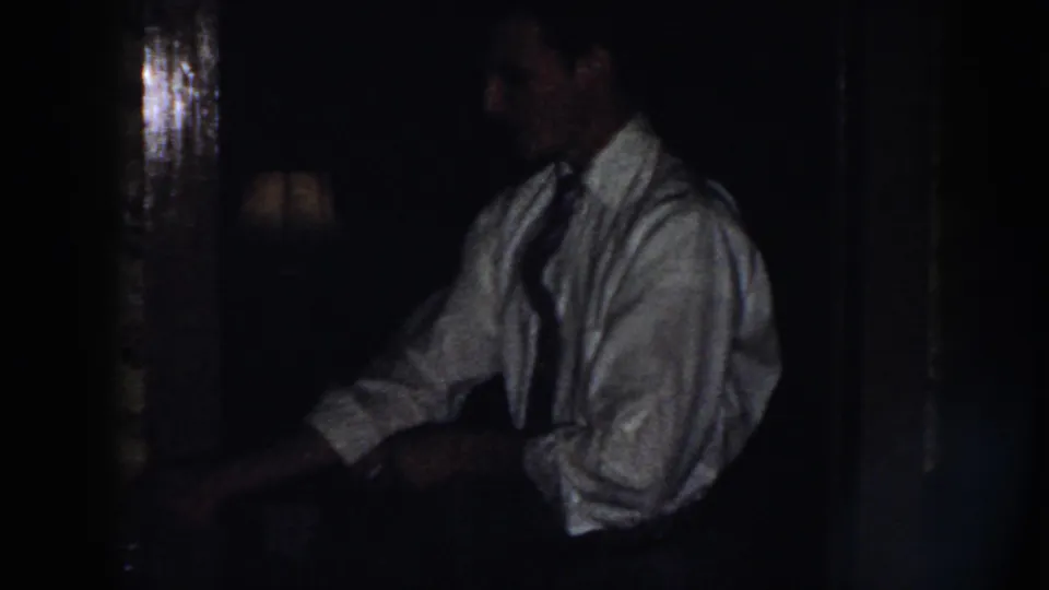 A low-light frame shows the man leaning slightly forward in a light shirt and tie, with his hands near a shirt cuff.