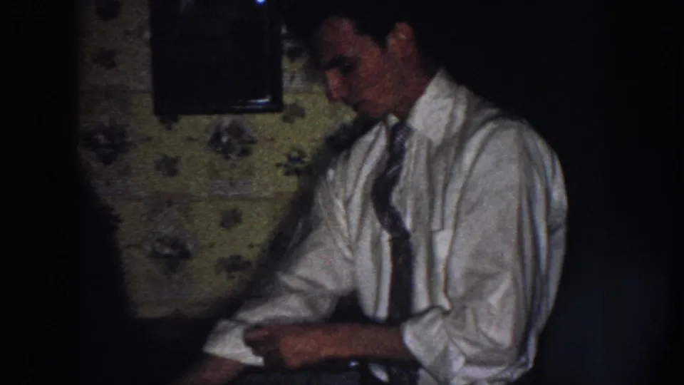 In the clearest frame, a man in a light dress shirt and tie adjusts a rolled sleeve beside floral wallpaper in a Nebraska home interior.