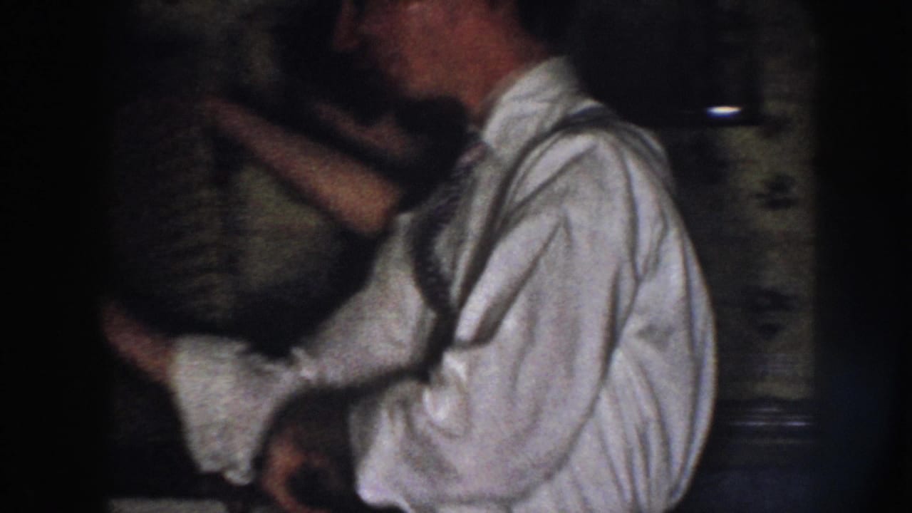 In a 1958 Nebraska home-movie frame, a man in a white shirt and tie rolls up his sleeve near a woman working at a dim kitchen sink.