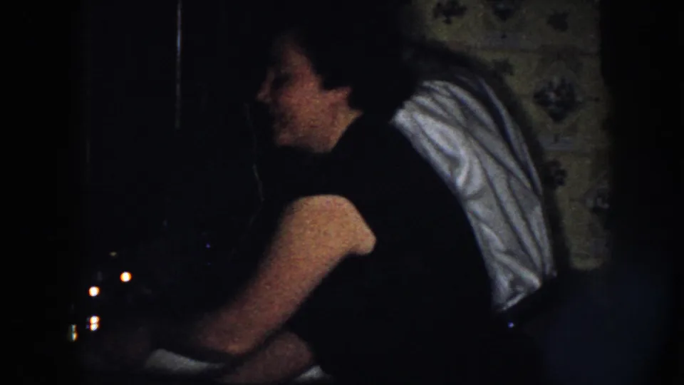 The woman smiles in profile as she leans near the sink, towel over her shoulder, in a dim 1958 Nebraska kitchen scene.