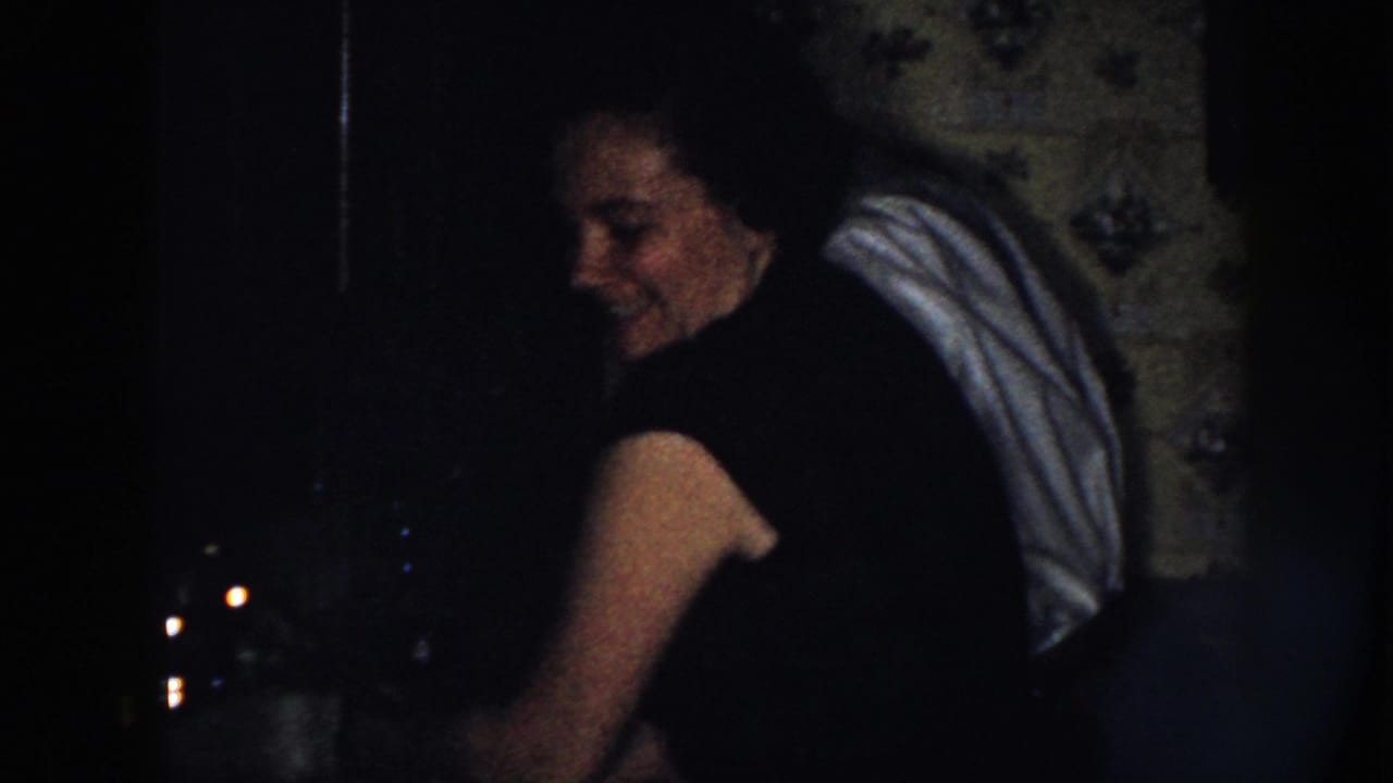 In 1958 Nebraska home-movie footage, a woman smiles toward the camera in a shadowed interior with a light cloth draped over her shoulder.
