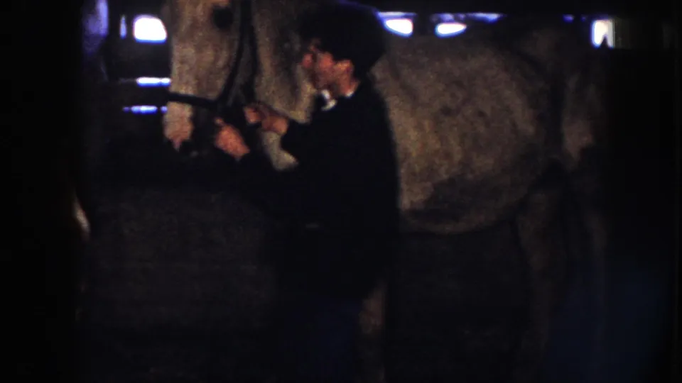 1958 Nebraska home-movie frame showing a person tending the head of a larger white horse inside a low-lit stable.
