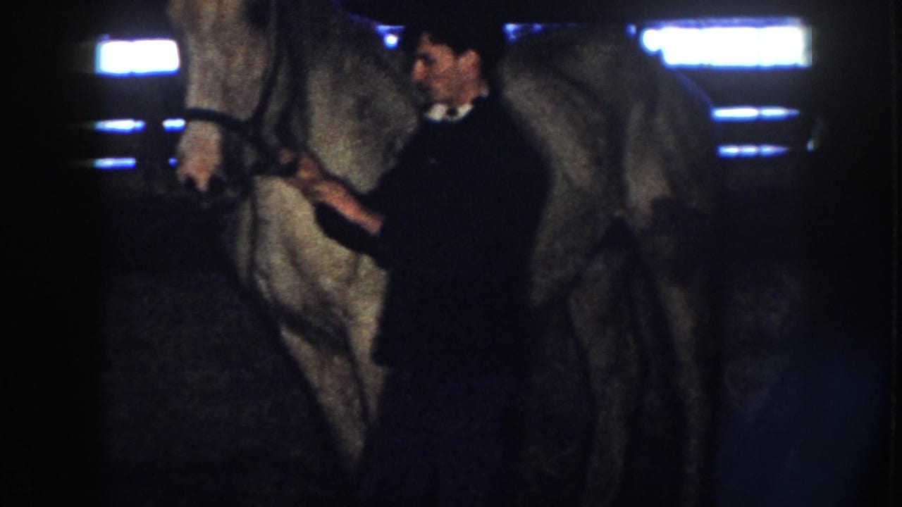 1958 Nebraska home-movie frame showing a man near a light-colored horse inside a dark stable with bright overhead lights.