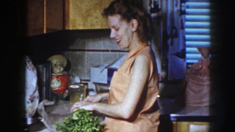 A smiling woman cuts fresh celery stalks in a retro kitchen, with a doll and containers visible behind her.