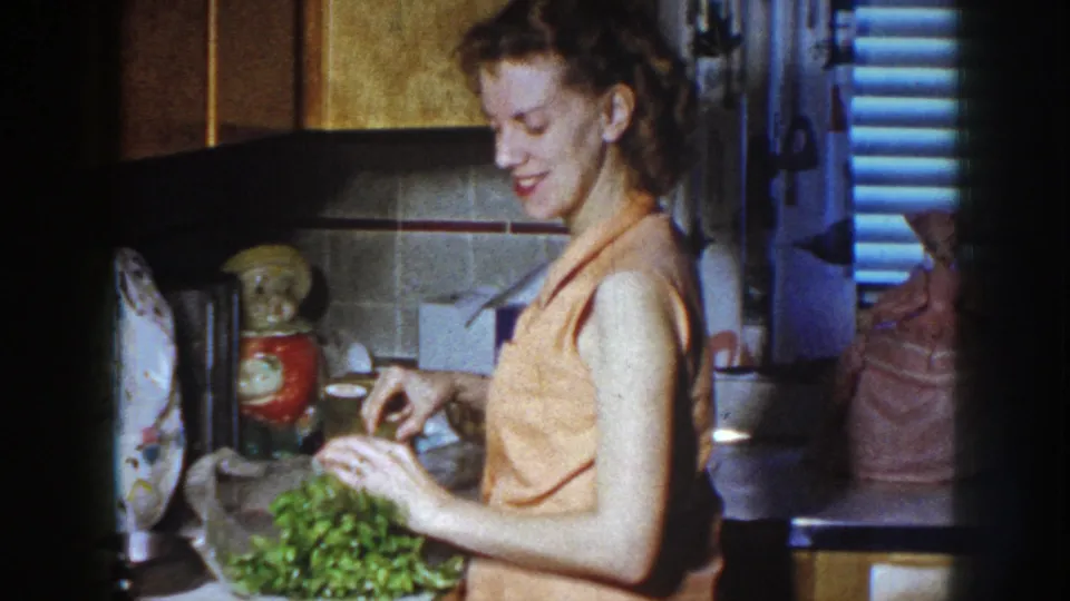 The woman holds celery at the kitchen counter, with mid-century kitchen objects and blue shutters visible in the background.