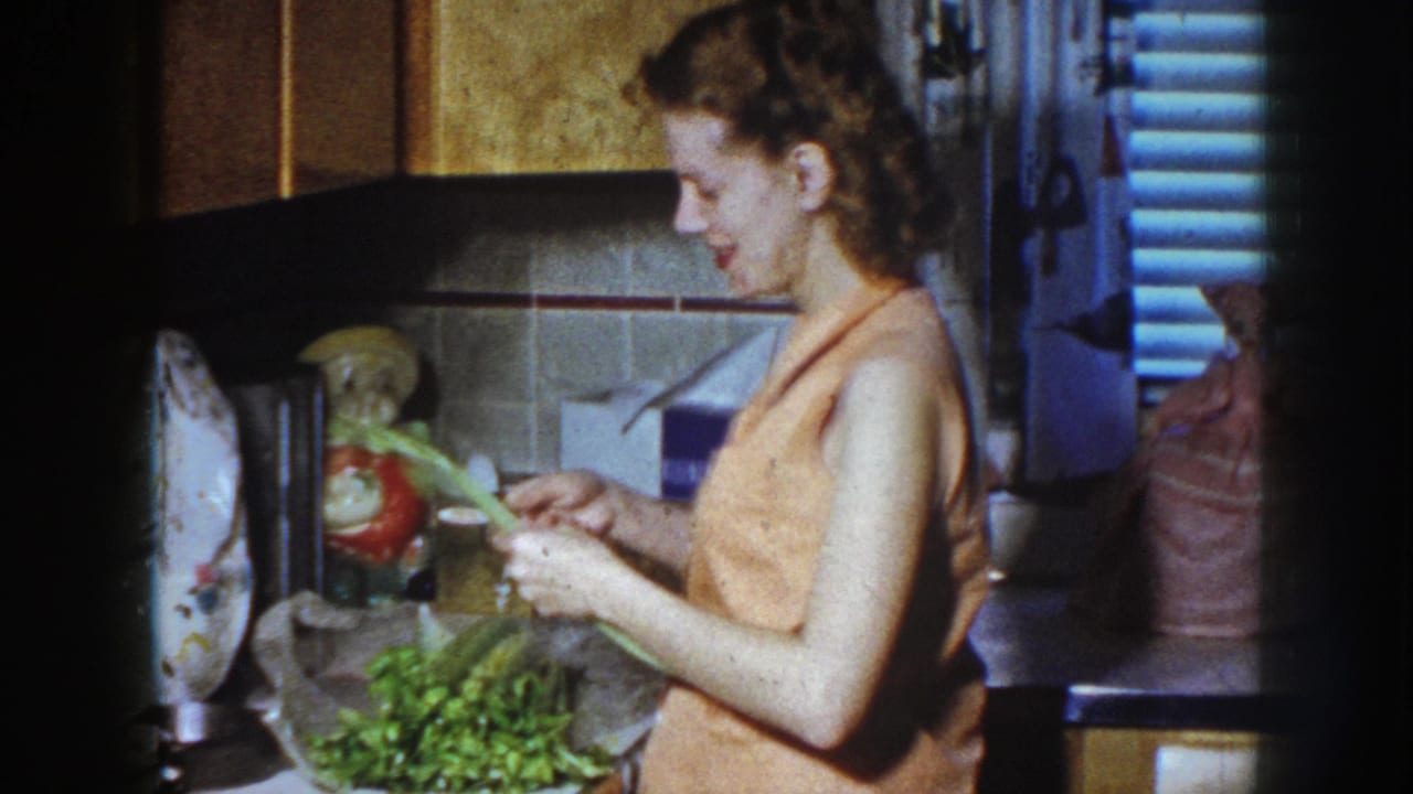 In a Nebraska 1958 home-movie frame, a woman in an orange sleeveless blouse trims green vegetables at a kitchen counter.