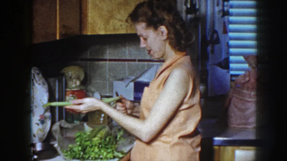 A woman in an orange blouse holds a celery stalk in a Nebraska kitchen, with vegetables gathered on the counter below.