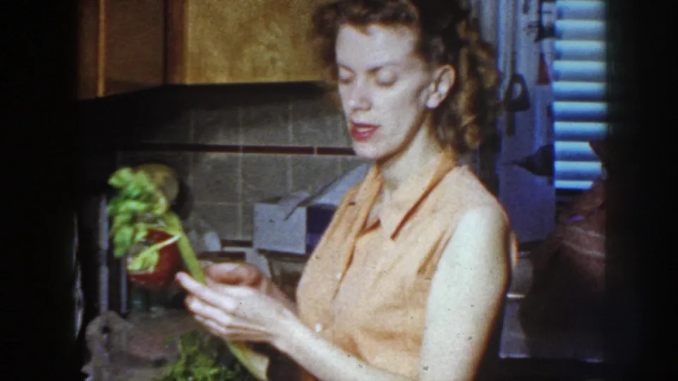 The woman holds leafy greens in a 1958 Nebraska kitchen, with countertop items and a tiled backsplash behind her.