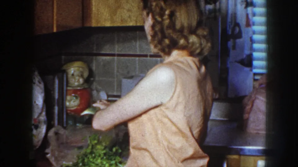 Adult woman in a 1958 Nebraska kitchen faces the counter near celery, a tiled wall, and a decorative ceramic figurine.