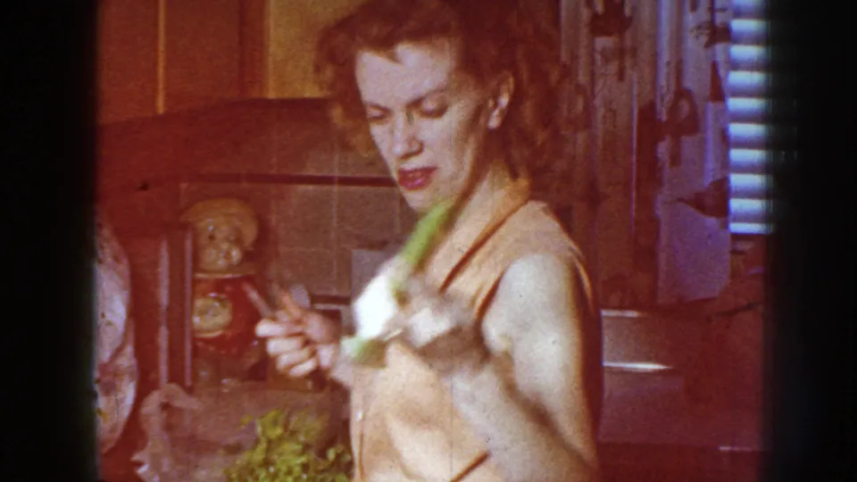 In a 1958 Nebraska home kitchen, the woman raises celery toward the camera with a knife in her other hand.