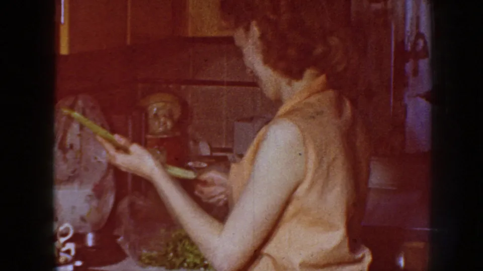 Adult woman in a sleeveless blouse holds celery near the counter in a 1958 Nebraska private kitchen interior.