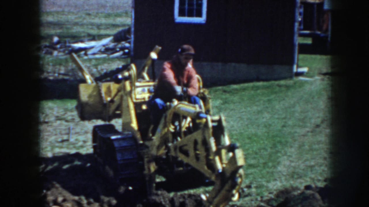 In 1958 Nebraska footage, a man in a red jacket and cap sits on a yellow tracked excavator beside a dark farm outbuilding.